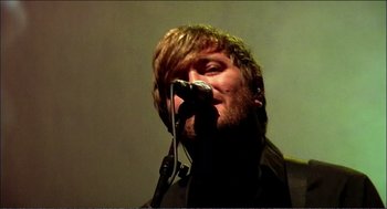 Movie still from “9 Songs” (2004), directed by Michael Winterbottom – A man holding a microphone up to his mouth; Close Up shot, Low angle