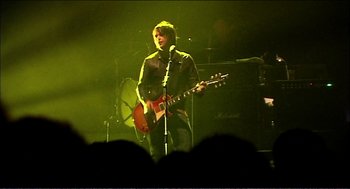 Movie still from “9 Songs” (2004), directed by Michael Winterbottom – A man with a guitar on stage in front of an audience; Medium shot, Low angle
