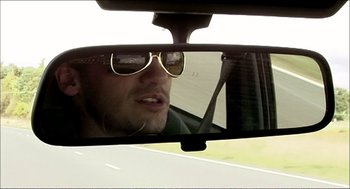 Movie still from “9 Songs” (2004), directed by Michael Winterbottom – A man wearing sunglasses looking in a rear view mirror of a car; Close Up shot, Over the shoulder angle