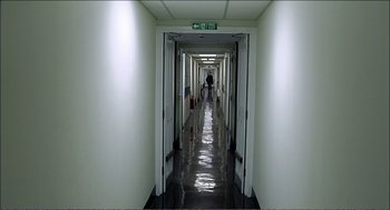 Movie still from “9 Songs” (2004), directed by Michael Winterbottom – A person walking down a long hallway in a building; Extreme Wide shot, High angle