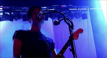 Movie still from “9 Songs” (2004), directed by Michael Winterbottom – A man with a microphone and a guitar; Medium shot, Low angle