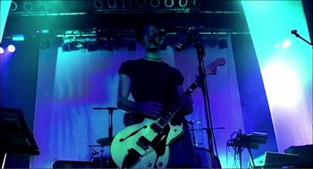 Movie still from “9 Songs” (2004), directed by Michael Winterbottom – A man playing a guitar in front of an audience; Medium shot, Low angle