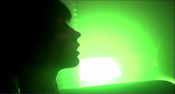 Movie still from “9 Songs” (2004), directed by Michael Winterbottom – A woman's face is lit up by a green light; Medium shot, Low angle