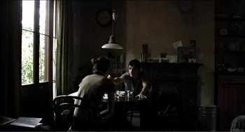 Movie still from “9 Songs” (2004), directed by Michael Winterbottom – Two people sitting at a table in a dark room; Medium shot, Over the shoulder angle