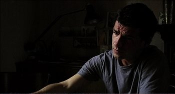 Movie still from “9 Songs” (2004), directed by Michael Winterbottom – A man sitting at a table in a dark room; Close Up shot, Low angle