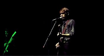 Movie still from “9 Songs” (2004), directed by Michael Winterbottom – A man holding a microphone while playing a guitar; Medium shot, Low angle