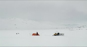 Movie still from “9 Songs” (2004), directed by Michael Winterbottom – A man standing in the snow next to an orange tent; Extreme Wide shot, High angle