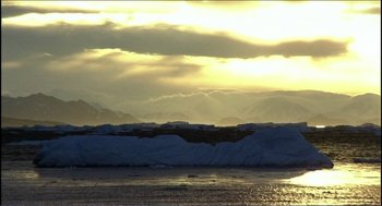 Movie still from “9 Songs” (2004), directed by Michael Winterbottom – The sun is setting over the ocean with a mountain range in the background; Extreme Wide shot, High angle