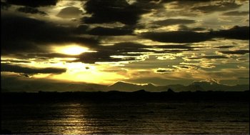 Movie still from “9 Songs” (2004), directed by Michael Winterbottom – The sun is setting over the ocean with mountains in the background; Extreme Wide shot, High angle