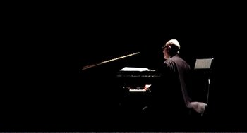 Movie still from “9 Songs” (2004), directed by Michael Winterbottom – A man sitting at a piano in the dark; Medium shot, Low angle