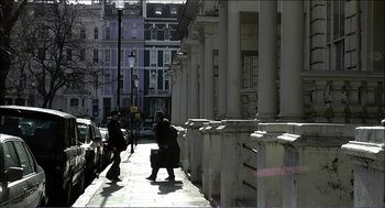 Movie still from “9 Songs” (2004), directed by Michael Winterbottom – Two people walking down a sidewalk near a building; Wide shot, Low angle