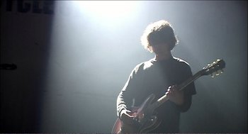 Movie still from “9 Songs” (2004), directed by Michael Winterbottom – A man playing a guitar in a dark room; Medium shot, Low angle