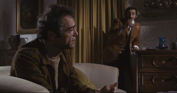 Movie still from “A Bay of Blood” (1971), directed by Mario Bava – A man sitting on a couch while another man takes a drink; Medium shot, Over the shoulder angle