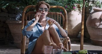 Movie still from “A Bigger Splash” (2015), directed by Luca Guadagnino – A woman sitting in a chair eating a donut; Medium shot, High angle