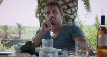 Movie still from “A Bigger Splash” (2015), directed by Luca Guadagnino – A man sitting at a table in front of a palm tree; Close Up shot, Over the shoulder angle
