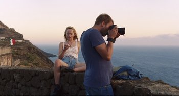 Movie still from “A Bigger Splash” (2015), directed by Luca Guadagnino – A man taking a picture of a woman sitting on a wall; Medium shot, Over the shoulder angle