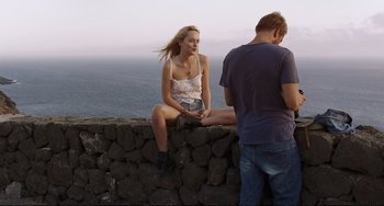 Movie still from “A Bigger Splash” (2015), directed by Luca Guadagnino – A man and a woman sitting on top of a stone wall; Wide shot, Over the shoulder angle