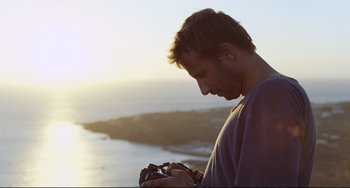 Movie still from “A Bigger Splash” (2015), directed by Luca Guadagnino – A man looking at a camera while standing by the ocean; Close Up shot, Low angle