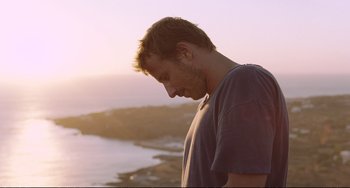 Movie still from “A Bigger Splash” (2015), directed by Luca Guadagnino – A man standing on top of a hill near a body of water; Close Up shot, Low angle