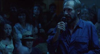 Movie still from “A Bigger Splash” (2015), directed by Luca Guadagnino – A man is holding a microphone in front of a group of people; Medium shot, Over the shoulder angle