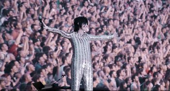 Movie still from “A Bigger Splash” (2015), directed by Luca Guadagnino – A woman in a silver outfit holding a microphone in front of a crowd; Wide shot, Low angle