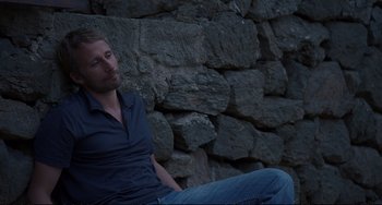 Movie still from “A Bigger Splash” (2015), directed by Luca Guadagnino – A man sitting on the ground in front of a stone wall; Medium shot, High angle