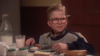 Movie still from “A Christmas Story” (1983), directed by Bob Clark – A young boy sitting at a dinner table with a plate of food in front of him; Medium shot, Low angle