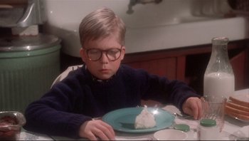 Movie still from “A Christmas Story” (1983), directed by Bob Clark – A young boy sitting at a table with a plate of cake on it; Medium shot, High angle