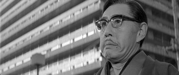 Movie still from “A Colt Is My Passport” (1967), directed by Takashi Nomura – An older man wearing glasses and looking at the camera; Close Up shot, Low angle