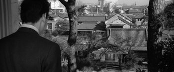 Movie still from “A Colt Is My Passport” (1967), directed by Takashi Nomura – A man standing next to a tree in front of some buildings; Medium shot, Low angle