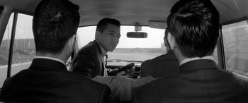 Movie still from “A Colt Is My Passport” (1967), directed by Takashi Nomura – A man in a suit sitting in the back of a car; Close Up shot, Over the shoulder angle
