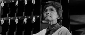 Movie still from “A Colt Is My Passport” (1967), directed by Takashi Nomura – An older woman standing in front of a wall with numbers; Close Up shot, Low angle