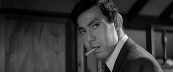 Movie still from “A Colt Is My Passport” (1967), directed by Takashi Nomura – A black - and - white photo of a man smoking a cigarette; Close Up shot, Low angle