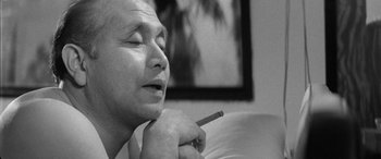 Movie still from “A Colt Is My Passport” (1967), directed by Takashi Nomura – An older man smoking a cigarette while sitting on a couch; Close Up shot, Low angle