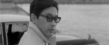 Movie still from “A Colt Is My Passport” (1967), directed by Takashi Nomura – Black and white photograph of a man wearing glasses; Close Up shot, Over the shoulder angle