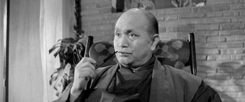 Movie still from “A Colt Is My Passport” (1967), directed by Takashi Nomura – An older man smoking a pipe while sitting in a chair; Close Up shot, Low angle