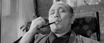 Movie still from “A Colt Is My Passport” (1967), directed by Takashi Nomura – An older man smoking a pipe while wearing a suit; Close Up shot, Low angle
