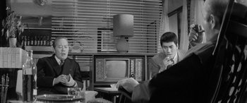Movie still from “A Colt Is My Passport” (1967), directed by Takashi Nomura – A man sitting at a table in front of a television; Medium shot, Over the shoulder angle