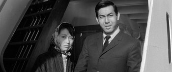 Movie still from “A Colt Is My Passport” (1967), directed by Takashi Nomura – An older man and a younger woman standing next to each other; Close Up shot, Low angle