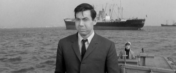 Movie still from “A Colt Is My Passport” (1967), directed by Takashi Nomura – Black and white photograph of a man wearing a suit and tie; Medium shot, Low angle