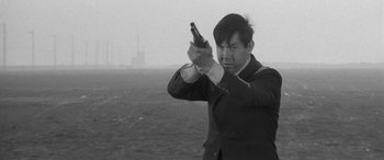 Movie still from “A Colt Is My Passport” (1967), directed by Takashi Nomura – A black and white photo of a man holding a gun; Medium shot, Over the shoulder angle