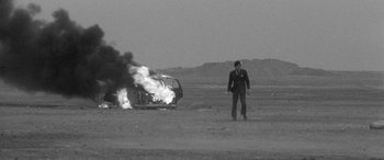 Movie still from “A Colt Is My Passport” (1967), directed by Takashi Nomura – A man standing in front of a car on fire; Wide shot, Over the shoulder angle