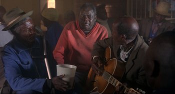 Movie still from “A Dry White Season” (1989), directed by Euzhan Palcy – Two men are sitting in a room and one of them is playing a guitar; Medium shot, Over the shoulder angle