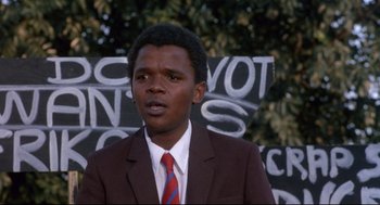 Movie still from “A Dry White Season” (1989), directed by Euzhan Palcy – A man in a brown suit and a red tie standing in front of a wall; Close Up shot, Low angle