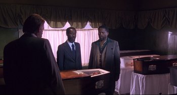 Movie still from “A Dry White Season” (1989), directed by Euzhan Palcy – A group of men standing around a wooden casket in a room; Medium shot, Over the shoulder angle
