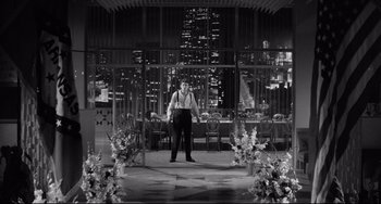 Movie still from “A Face in the Crowd” (1957), directed by Elia Kazan – A black and white photo of a man standing in front of a table; Wide shot, High angle