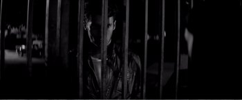 Movie still from “A Girl Walks Home Alone at Night” (2014), directed by Ana Lily Amirpour – A man standing behind a metal fence at night; Medium shot, Low angle