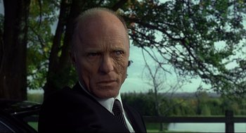 Movie still from “A History of Violence” (2005), directed by David Cronenberg – An older man wearing a suit and tie in front of trees; Close Up shot, Low angle