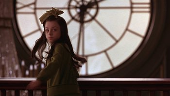Movie still from “A Little Princess” (1995), directed by Alfonso Cuarón – A young girl with a bow on her head standing in front of a clock; Medium shot, Over the shoulder angle
