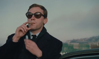 Movie still from “A Man and a Woman” (1966), directed by Claude Lelouch – A man in a suit and sunglasses smoking a cigarette; Medium shot, Low angle